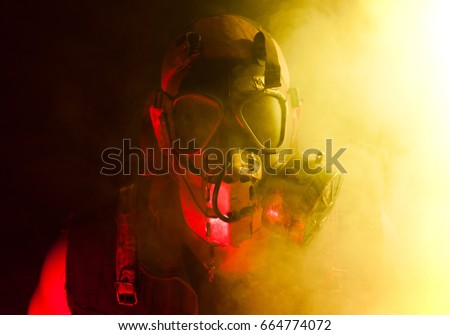 A creepy guy with post apocalyptic vibes wearing a gas mask in a smoke or fog cloud that's colored bright yellow while being lit with a red light from other side.