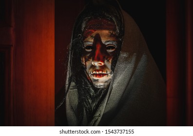 Creepy halloween witch with black eyes, scars on her face in a hood stands near opened doors in the night. Portrait of a scary creature from another world
