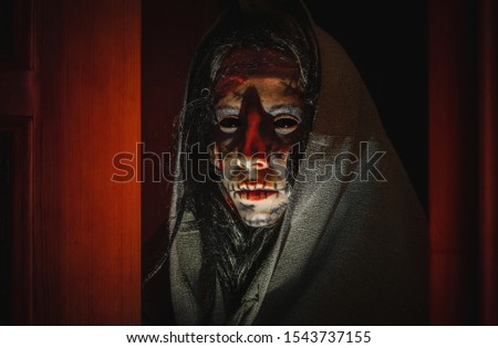 Creepy halloween witch with black eyes, scars on her face in a hood stands near opened doors in the night. Portrait of a scary creature from another world