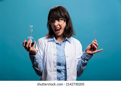 Creepy looking biochemistry specialist holding glass vials filled with chemical liquid substance samples on blue background. Microbiology crazy and lunatic looking expert having chemistry jars.