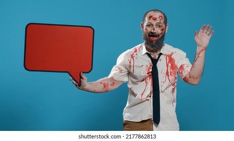 Creepy Mindless Zombie Holding Red Cardboard Speech Bubble On Blue Background. Frightening Graphic Brain Dead Monster Having Carton Banner While Waving At Camera. Studio Shot.