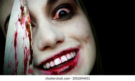 A Creepy Woman Covered In Blood Stains Holds A Knife And Looks At The Camera, Isolated On Black Background. Crazy Smile. Halloween Or Horror Theme. 