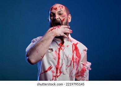 Creepy Zombie Biting Hand With Scars And Bloody Wounds, Standing In Studio. Apocalyptic Frightening Corpse And Brain Eating Villain With Deadly Horror Face And Aggressive Sinister Eyes.