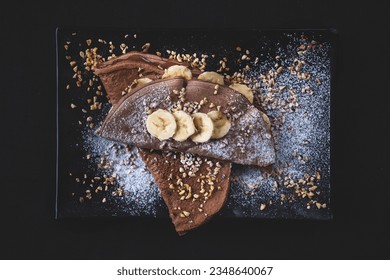 Crepes (thin pancakes) in chocolate, with peanut butter, slices of banana, chopped hazelnuts on the black ceramic plate. Morning breakfast, snack or homemade dessert. Black background. View from above - Powered by Shutterstock