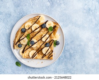 Crepes (thin pancakes, traditional russian dessert blini) with fresh berries, banana slices, melted chocolate and green mint. Maslenitsa concept. Tasty morning breakfast. Blue background, copy space.