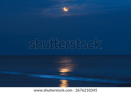 A crescent moon casts a golden reflection across the still water under a deep blue night sky.