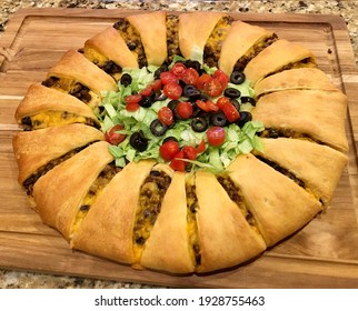 Crescent Rolls Taco Ring on a Wood Cutting Board, made of hamburger, cheese, tomatoes, lettuce, and black olives