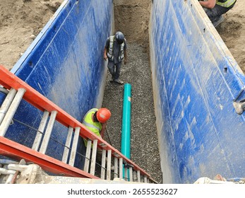 Crew inspects initial SDR35 segment for alignment and surface defects while awaiting next pipe delivery inside shored trench for sanitary main install.