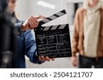 Crew member holding film clapper while preparing for a movie scene on a film set. Blurred background includes another crew member standing, adding depth to film production scene