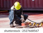 A crew member kneels on the deck of a merchant ship, using a pneumatic tool to remove rust. Focused on preparing the surface for painting and maintenance, ensuring the ship