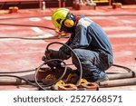 A crew member kneels on the deck of a merchant ship, using a pneumatic tool to remove rust. Focused on preparing the surface for painting and maintenance, ensuring the ship