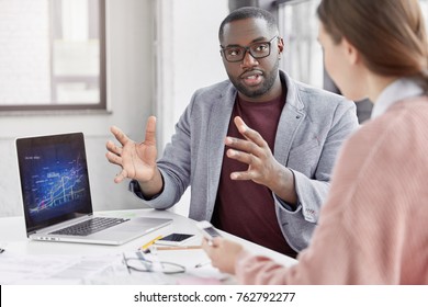 Crew Of Talented Young Employees Explain Ideas During Working Meeting. Black Male Employer Shows Financial Presentation, Analyze Graphics, Gestures With Hands, Look Attentively At Interloutor