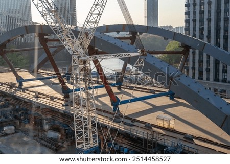 Crews work on the construction of a bridge over a river, with cranes and steel beams prominent in the bustling urban environment