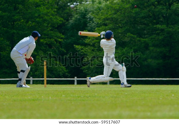 A cricket
batsman playing a pull shot towards the boundary in a cricket match
while the catcher looks on.