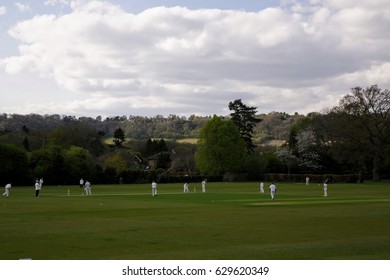 Cricket Match, Oxted, UK. Men Dressed In Traditional Whites Dotted About An English Village Green, Clouds Above.