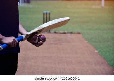 Cricketer Holds Old Red Leather Cricket Ball And Wooden Cricket Bat In Hands On Pitch, Soft And Selective Focus, Blurred Cricket Field Background.