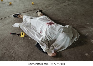 Crime Scene With Body Of Shot Dead Man Covered With White Sheet On Asphalt Surrounded By Knife With Blood And Evidence Numbers