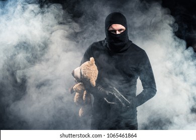 Criminal in balaclava holding gun and teddy bear in clouds of smoke