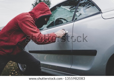 Criminal in black balaclava and hoodie opens somebody's vehicle with skeleton key. Car breaking by unknown male with hidden face. Young man picks automobile lock to get inside. Forcing the car door.