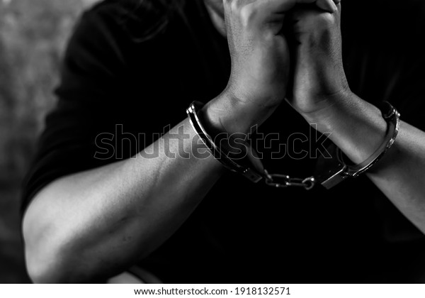 Criminal concept Bad guy was cuffed and under\
arrest by policeman and detain prisoner in jail Detainees get\
hopeless and stressed in dirty prison Young prisoner sit in dark\
room Black and white\
photo