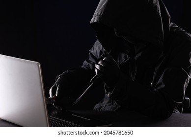 Criminal hacker in a hood in black clothes and balaclava destroys a laptop with tools, a screwdriver and pliers on a black background. - Powered by Shutterstock