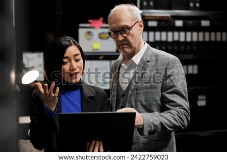 Criminal private investigation team in agency cabinet room comparing criminology suspect case file details on laptop. Asian private investigator and senior detective in archival repository office