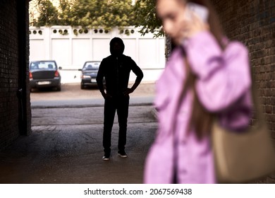 Criminal stalking a woman alone in dark street alley. Female is talking on phone. Unrecognizable man is hiding in the shadows. Woman Doesn't Suspecting That She Is Being Followed By Criminal Bandit