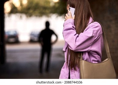 Criminal stalking woman, commiting crime while victim was walking alone, talking on phone in dark street. Caucasian young woman is looking back, afraid of male stranger person in the background - Shutterstock ID 2088744106