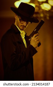 Criminal World, Mafia. Portrait Of A Cunning Brunet Man In Black Coat And A Hat Standing In Dark Room, Lit By The Ray Of Light, With A Gun In His Hand. Retro Noir Style. 