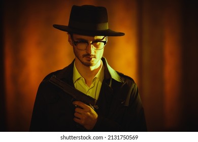 Criminal World, Mafia. Portrait Of A Thoughtful Brunet Man In Black Coat And A Hat Standing In Dark Room, Lit By A Ray Of Light, With A Gun In His Hand. Retro Noir Style. 