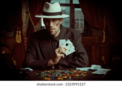 The criminal world of the mafia. A tough gangster man in an elegant black suit and a white hat sits in a luxury apartment and plays cards. Retro style. Detective movie.