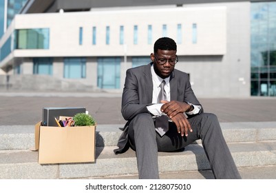 Crisis And Unemployment Concept. Upset Black Office Worker Sitting With Box Of Personal Stuff On Stairs Of Office Center, Got Fired, Man Losing His Job