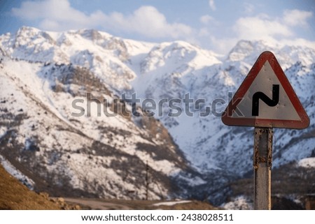 The crisp air of an alpine road is marked by a cautionary traffic sign, warning of the curving path ahead. Behind, the grandeur of snow-dusted mountains looms, signifying the beauty and perils of moun