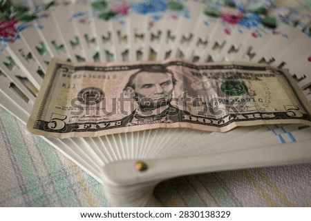 A crisp five dollar bill lying across an open white decorated hand fan on a tablecloth surface.