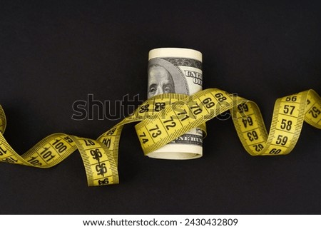 A crisp hundred dollar bill is tightly rolled and encircled by a yellow measuring tape with visible numbers, suggesting themes of financial measurements and value assessment against a dark