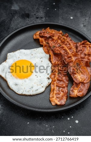 Crispy bacon and bright fried egg on black plate over dark textured background, perfect for breakfast, keto meals and gourmet food themes