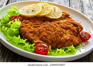 Crispy breaded seared chicken cutlet with lemon slices and fresh vegetables on wooden table 