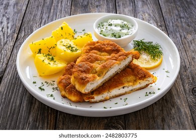 Crispy breaded seared chicken cutlet with boiled potatoes and cucumbers in cream served on white plate on wooden table