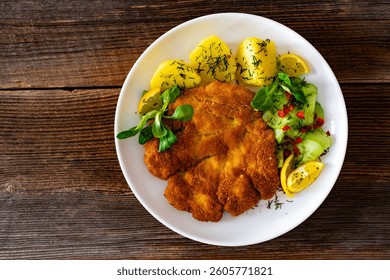  Crispy breaded seared chicken cutlet with boiled potatoes and fresh cucumbers served on wooden table