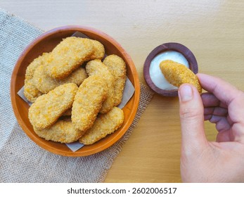 Crispy and crunchy fried chicken nuggets served on a wooden plate. And a hand is dipping a fried chicken nugget into a small wooden bowl filled with mayonnaise sauce. - Powered by Shutterstock - Get 15% off with code: PIKWIZARD15