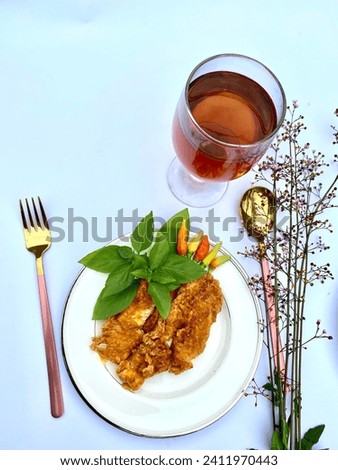Crispy deep fried chicken breast on the white plate garnished with fresh basil leafs and colorful chilly,  served with ice tea, fox and spoon, tiny flower bud as decoration