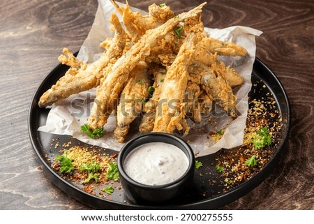 crispy, deep-fried small fish, likely smelt or whitebait, served as an appetizer with a creamy white dipping sauce on a black plate. The dish is garnished with herbs and spices.