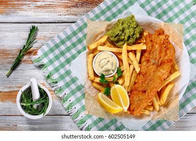 crispy fish and chips - fried cod, french fries, lemon slices, tartar sauce and mashed peas on plate on paper on old wooden table with rosemary in mortar, authentic recipe, view from above