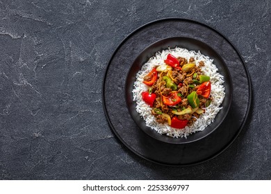 crispy ground beef with stir-fried veggies and a sticky-sweet orange sauce topped on rice in black bowl on concrete table, horizontal view from above, flat lay, copy space