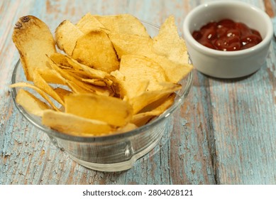 Crispy kettle cooked potato chips filling a glass bowl, served next to a small dish of red ketchup on a rustic wooden table