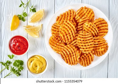 Crispy Potato Waffles Fries, Wavy, Crinkle Cut, Criss Cross Fries on a white plate on a wooden table with mustard and tomato sauce dipping, view from above, flat lay