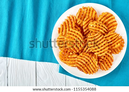 Crispy Potato Waffles Fries, Wavy, Crinkle Cut, Criss Cross Fries on a white plate on a textile table mat on a wooden table, view from above, flat lay 