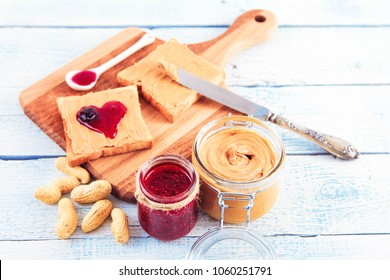 
Crispy Toasts With Peanut Butter And A Heart Of Jam Lie On A Wooden Board On A White Wooden Background. A Jar Of Raspberry Jam. Useful, Healthy And Tasty Breakfast.