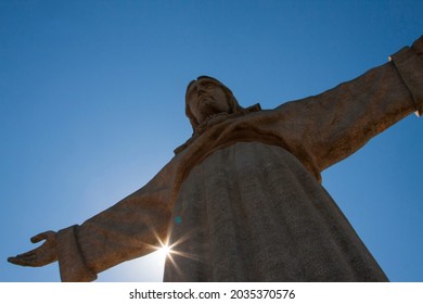 Cristo Rei monument (Christ king) in Lisbon, Portugal, against a blue sky and sunrays peaking under one of its arms.
