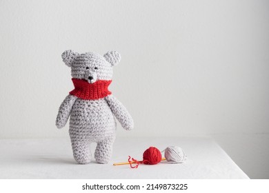 Crochet knitting cute gray, white, polar teddy bear with a red scarf stands on the table, homemade amigurumi with knitting hook and yarn